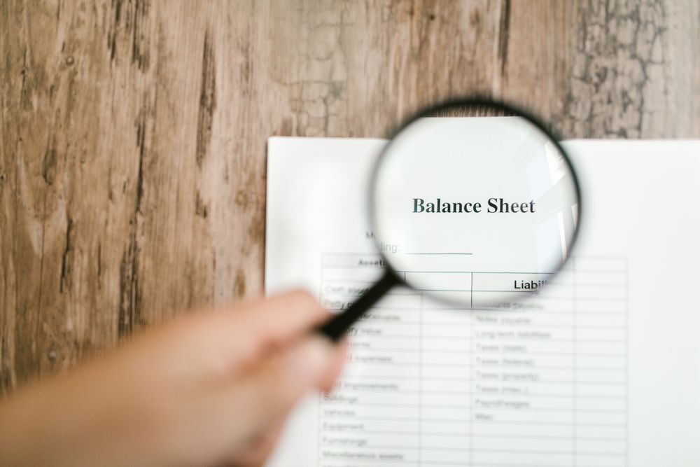 A hand holds a magnifying glass over a printed financial balance sheet on a wooden desk
