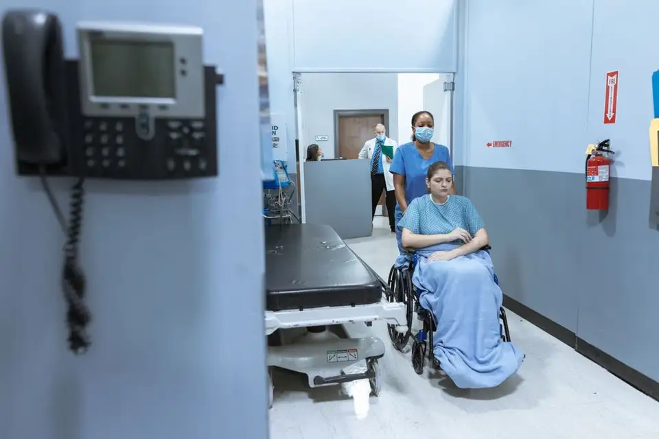 A nurse guides a patient in a wheelchair down a bright hospital corridor.