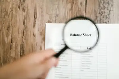 A hand holds a magnifying glass over a printed financial balance sheet on a wooden desk
