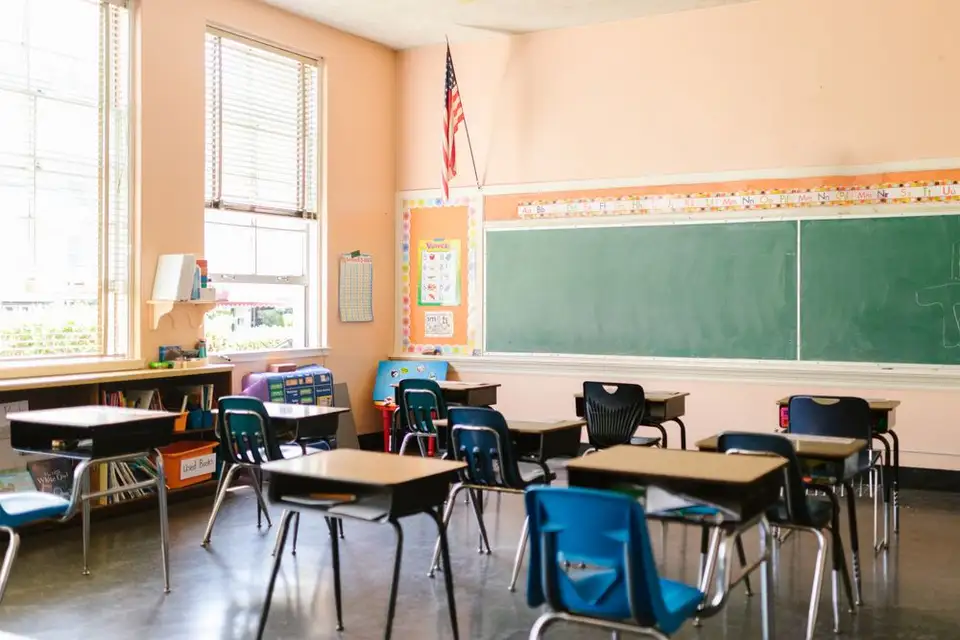 Rows of empty desks in a sunlit classroom beneath an American flag.
