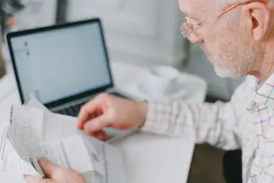 An elderly man in glasses reading a stack of documents at home